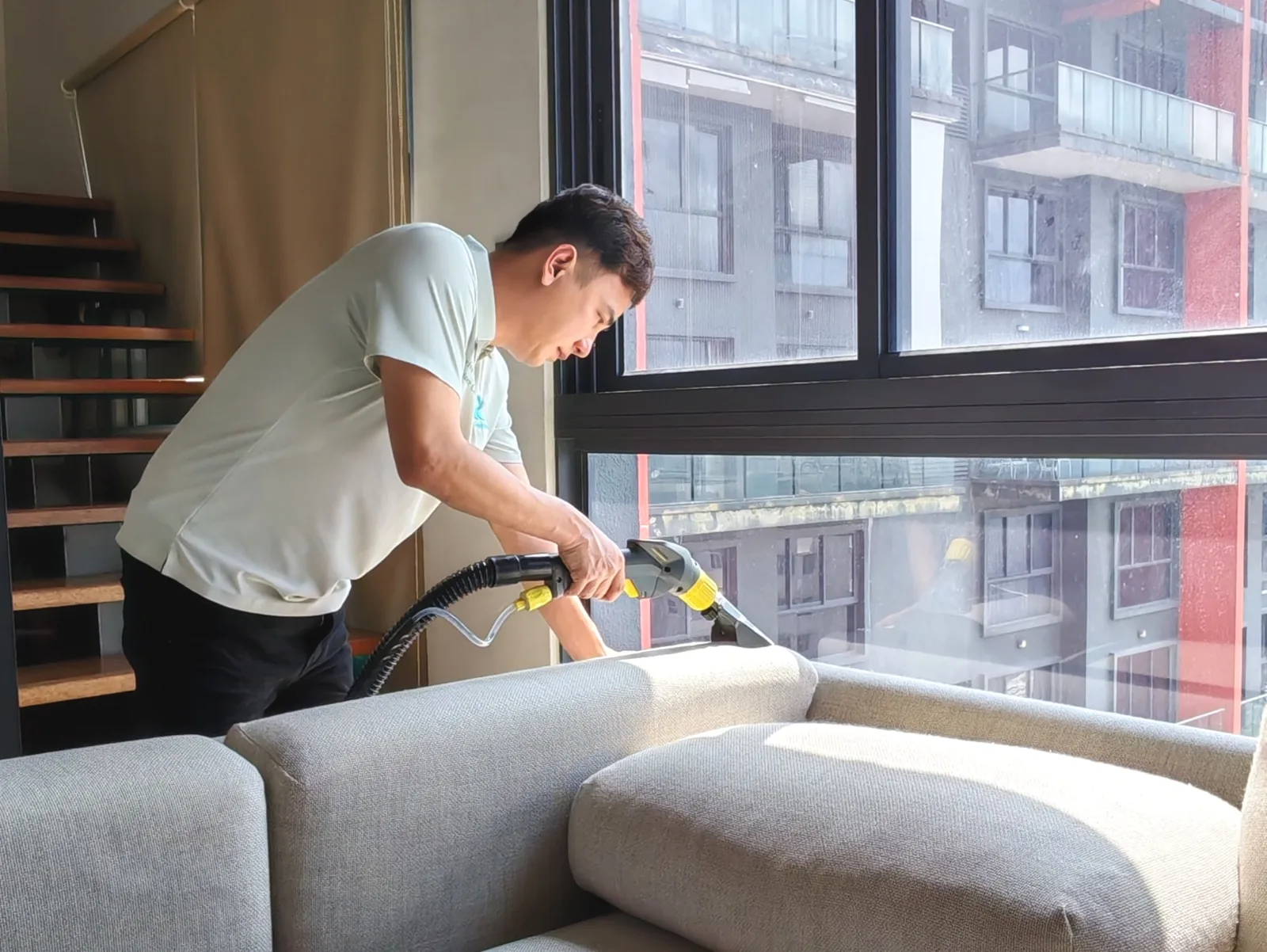 Technician cleaning a grey sofa seat beside a window